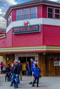 People walk past the entrance of the Red Dog Saloon, a historic red-painted bar and restaurant in downtown Juneau, Alaska. The building features rustic wood accents, a large sign with a red dog logo, and an inviting open doorway with visitors entering and exiting.